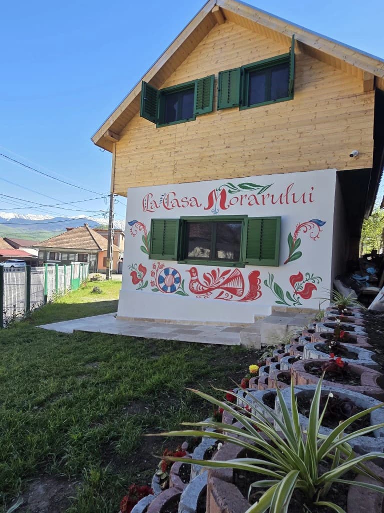 Fațada La Casa Morarului din Cârțișoara: nume pictat, motive populare roșii și albastre, etaj din lemn cu obloane verzi și creste Făgăraș în zare